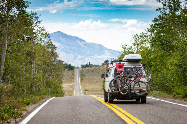 Family camper van with bicycles mounted on the rear driving down a scenic mountain highway lined with trees, with a winding road and mountain peaks visible in the distance under a blue sky.