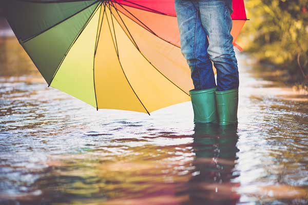 Child standing in large puddle with green boots and rainbow umbrella