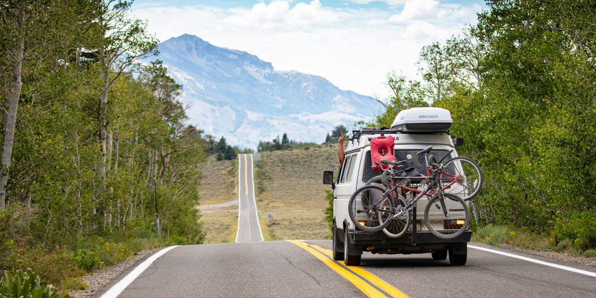 A camper van with bicycles mounted on the back drives along a scenic two-lane road lined with trees, with mountains rising in the background under a partly cloudy sky.